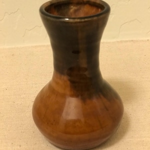 May include: A small, handcrafted ceramic vase with a bulbous base and a narrow neck. The vase features a gradient of colors, transitioning from a dark brown at the top to a warm, earthy brown at the base. The vase is approximately 4 inches tall.