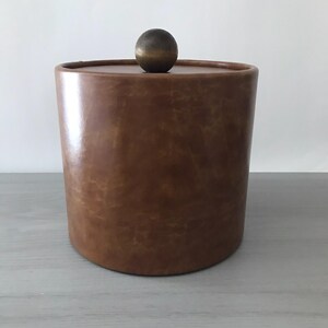 May include: A brown leather-like ice bucket with a round lid and a small, spherical handle. The cylindrical bucket has a smooth, glossy finish. The lid is a darker shade of brown than the bucket. The handle is a bronze color.