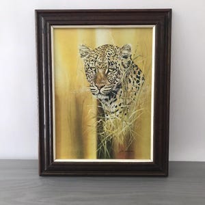 May include: A framed painting of a leopard peering through tall grass. The leopard has a spotted coat and is set against a yellow and brown background. The frame is dark brown.