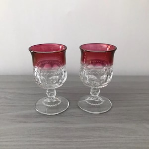 May include: Two clear glass goblets with a ruby red rim and a decorative etched pattern. The goblets have a gold rim and a clear stem and base. The goblets are on a gray surface.