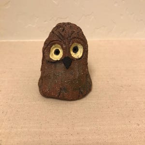 May include: A handcrafted, brown ceramic owl figurine with large, round yellow eyes and a black beak. The owl has a textured surface and a slightly rough appearance, suggesting it is handmade. The owl is a decorative item.