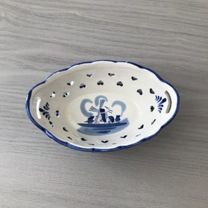 May include: Oval-shaped white ceramic dish with blue trim and a windmill scene. The dish has heart-shaped cutouts around the rim and decorative blue floral accents. A charming piece for serving or display.