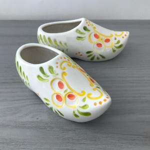 May include: Two white ceramic clogs with hand-painted floral designs in yellow, orange, and green. The clogs are a traditional Dutch design, with a rounded toe and open top. The clogs are displayed on a gray surface.