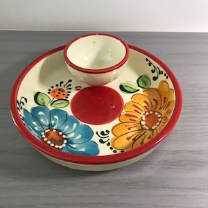 May include: A ceramic serving dish with a small bowl, featuring a cream-colored base and a red rim. The dish is decorated with blue and yellow floral designs and a red center. The small bowl is cream-colored with a red rim.