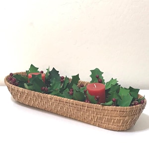May include: An elongated, woven tan basket filled with artificial green holly leaves and small red berries. Two cylindrical red pillar candles are nestled among the greenery, creating a decorative arrangement for a tabletop display.