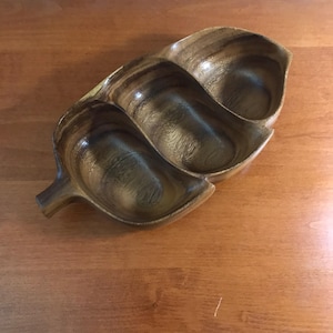 May include: A wooden leaf-shaped serving dish with three compartments. The dish is made of dark brown wood with a natural grain pattern. The dish is designed for serving snacks or appetizers.