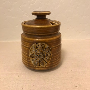 May include: A brown ceramic jar with a lid and a decorative floral design. The jar has horizontal ridges and a small handle on the lid. The floral design is in a circular shape on the front of the jar.