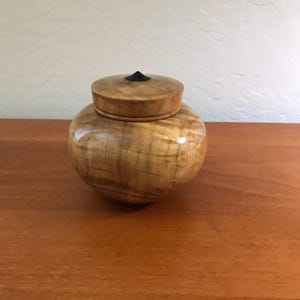 May include: A handcrafted wooden jar with a lid. The jar is spherical with a light brown finish and visible wood grain. The lid is circular with a small black knob on top. The jar sits on a wooden surface.