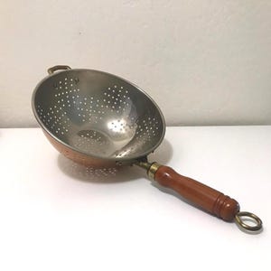 May include: A round metal colander with a polished silver-toned interior and a copper-toned exterior. The kitchen tool features a long, smooth brown wooden handle with a brass-colored metal ferrule and a circular hanging loop at the end.