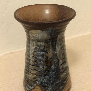 May include: A handmade ceramic vase with a wide, brown rim and a textured, cylindrical body. The vase features a blend of blue, gray, and brown hues, creating a unique, organic pattern. The interior is a darker brown.