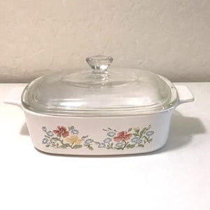 May include: A rectangular white casserole dish with integrated side handles and a clear glass lid featuring a knob handle. The exterior of the white dish is decorated with a floral pattern consisting of red, yellow, and blue flowers with green leaves.