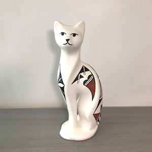 May include: A white ceramic cat figurine with black, brown, and white geometric patterns. The cat is in a seated position with its ears perked up. The figurine has a minimalist design.
