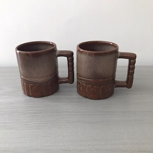 May include: Two brown ceramic mugs with handles. The mugs have a textured, speckled finish and a decorative band near the base. The handles are rectangular and attached to the side of the mugs. The mugs are designed for hot beverages.