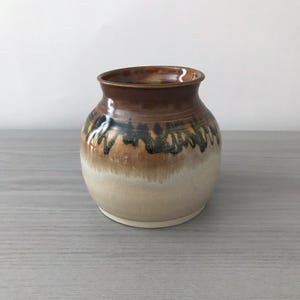 May include: A handcrafted ceramic vase with a rounded body and a wide opening. The vase features a gradient of colors, transitioning from a creamy white base to a brown rim, with a band of brown and black accents. The vase is suitable for home decor.