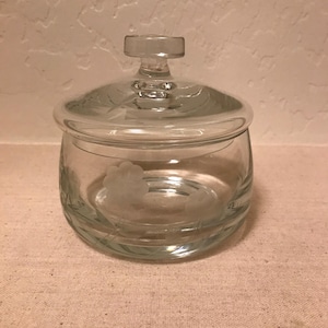 May include: A clear glass jar with a lid. The jar has a round shape with etched floral designs around the base and on the lid. The lid has a small handle. The jar is sitting on a neutral surface.