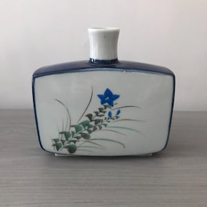 May include: A rectangular ceramic vase with a white body, a dark blue rim, and a white neck. The vase is decorated with a blue floral design and green leaves. The vase is sitting on a gray surface.