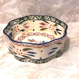 May include: A ceramic bowl with a scalloped edge, featuring a hand-painted floral design in red, blue, and green on a cream background. The bowl has a blue rim and a decorative border.