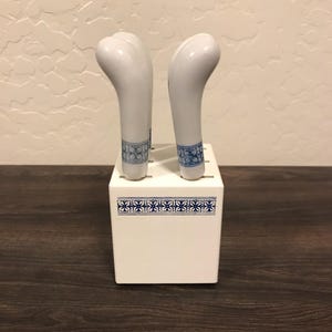 May include: Two white ceramic knives with curved handles, displayed in a white block. The handles have a blue floral pattern near the base. The block also features a blue floral design.
