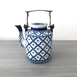 May include: A blue and white ceramic teapot with a brass handle. The teapot has a cylindrical shape with a spout and a lid. The surface is decorated with a repeating floral pattern. The handle is attached to the top of the teapot with metal brackets.