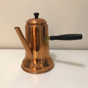 May include: A polished copper coffee pot with a cylindrical body, a long angled spout, and a black handle with horizontal ridges. The pot features a matching copper lid topped with a small black knob, showcasing a reflective metallic finish.