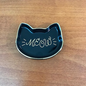May include: A black ceramic cat-shaped dish with gold trim. The word "MEOW" is written in gold script across the center. The dish is on a wooden surface.