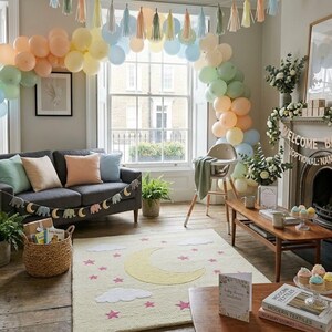 May include: A room decorated for a baby shower with pastel balloons, a gray sofa, and a rug with a moon and star design. A high chair and a wooden coffee table are also present. A banner reads "Welcome Baby".