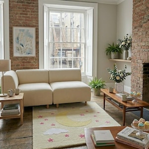 May include: A cozy living room scene featuring a cream-colored sectional sofa, a wooden coffee table, and a rug with a moon and star design. The room has a brick wall, a large window, and a painting on the wall. Plants and decorative items add to the inviting atmosphere.