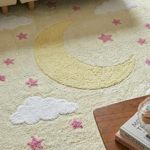 May include: A cream-colored rug with a yellow crescent moon, white clouds, and pink stars. The rug has a soft, textured surface. The design is whimsical and suitable for a nursery or children's room.