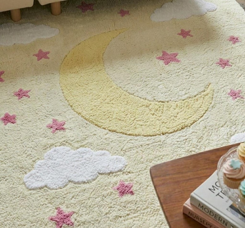 May include: A cream-colored rug with a yellow crescent moon, white clouds, and pink stars. The rug has a soft, textured surface. The design is whimsical and suitable for a nursery or children's room.