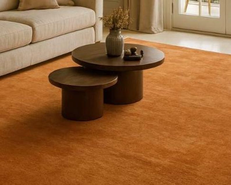 Puede incluir: Una escena de sal&oacute;n con una alfombra de color naranja. Dos mesas de centro redondas de color marr&oacute;n oscuro de diferentes tama&ntilde;os est&aacute;n centradas en la alfombra. Un sof&aacute; beige es parcialmente visible a la izquierda, y un jarr&oacute;n con flores secas.