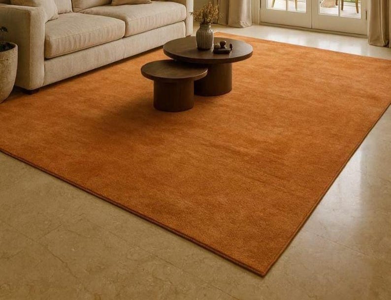 Puede incluir: Una alfombra rectangular naranja con una superficie texturizada. La alfombra est&aacute; colocada sobre un suelo de color claro, con un sof&aacute; beige y dos mesas de centro de madera marr&oacute;n en el fondo. La alfombra a&ntilde;ade calidez al espacio habitable.