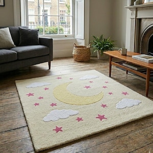 May include: A rectangular cream-colored rug with a whimsical design. It features a large yellow crescent moon, white fluffy clouds, and pink stars scattered across the surface. The rug is placed on a wooden floor in a living room setting.
