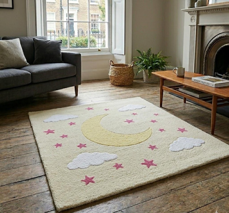 May include: A rectangular cream-colored rug with a whimsical design. It features a large yellow crescent moon, white fluffy clouds, and pink stars scattered across the surface. The rug is placed on a wooden floor in a living room setting.