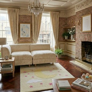 May include: A cozy living room with a cream-colored sectional sofa, a rug with a moon and star design, and a brick fireplace. The room is decorated with gold-framed artwork, a chandelier, and cream-colored curtains. A small wooden side table holds books and a lamp.