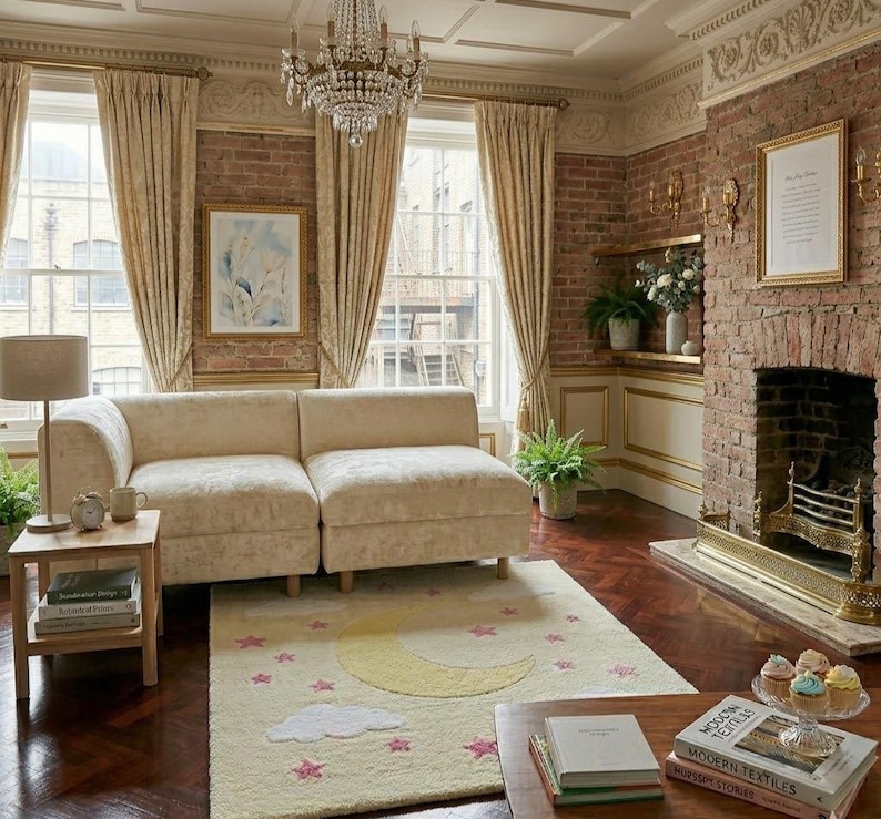 May include: A cozy living room with a cream-colored sectional sofa, a rug with a moon and star design, and a brick fireplace. The room is decorated with gold-framed artwork, a chandelier, and cream-colored curtains. A small wooden side table holds books and a lamp.