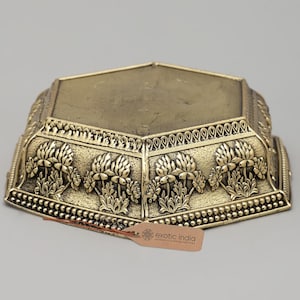 May include: A gold-colored, hexagonal decorative stand with floral relief carvings. The stand has a flat top and ornate detailing around the edges. The tag reads "exotic india authentic curated heritage".