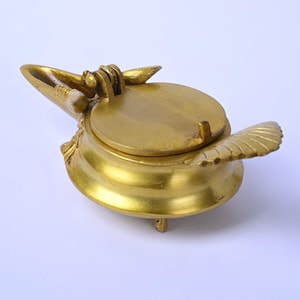 May include: A gold-coloured, ornate container with a hinged lid and a decorative wing-like element. The container has a round base and is elevated by small feet. The text "exotic india" is visible in the upper left corner.