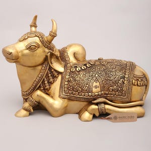 May include: A golden brass sculpture of a reclining cow, featuring intricate embossed details on its body and decorative elements. The sculpture has horns and is adorned with ornate jewelry. The cow is a symbol of heritage and culture.