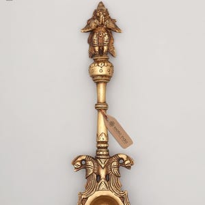 May include: A gold-colored, ornate oil lamp with a detailed design. The lamp features a figure at the top, a long stem, and a bowl-shaped base with a decorative border. The tag reads "exotic india".