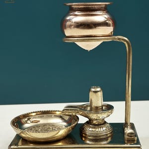 Könnte beinhalten: Ein religiöses Artefakt aus Messing mit einem kleinen Topf, der über einem Lingam und einer dekorativen Schale aufgehängt ist. Die Struktur besteht aus Messing und steht auf einem rechteckigen Sockel. Der Hintergrund ist türkis.