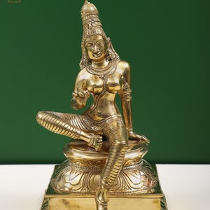 Puede incluir: Una estatua dorada de latón de una deidad sentada, adornada con intrincadas joyas y un tocado alto. La figura está sentada sobre un pedestal detallado, con una expresión serena y una pose elegante. La estatua es una representación del arte y la cultura indios.