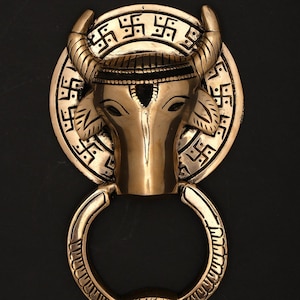 May include: A brass door knocker featuring a bull's head with curved horns, a circular backplate with etched symbols, and a ring-shaped handle. The design is detailed and ornate, with a vintage aesthetic.