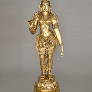 May include: A gold-colored statue of a female figure, adorned with intricate details. The statue features a detailed crown, jewelry, and ornate clothing. The figure stands on a decorative base. The image includes the text "exotic india authentic curated heritage".