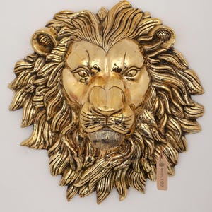 Pode incluir: Escultura de parede com cabeça de leão dourada com juba e traços faciais detalhados. A escultura tem uma etiqueta que diz "exotic india". O rosto do leão está centrado, com uma superfície texturizada e uma expressão régia. A escultura é provavelmente de metal.