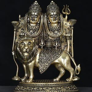 Può includere: Una scultura in ottone dettagliata raffigurante due figure con copricapi e gioielli ornati, sedute su un leone. La scultura presenta dettagli intricati ed è ambientata su uno sfondo scuro. Il testo "exotic india" è in alto.