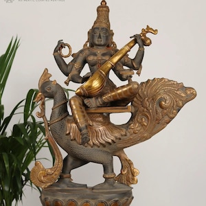 May include: A bronze statue of a deity playing a stringed instrument, seated on a peacock. The statue has multiple arms and is adorned with gold accents. The base is intricately detailed with floral patterns. The statue is from Exotic India.