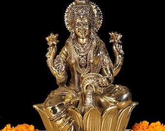 May include: A golden Lakshmi statue seated on a lotus flower, adorned with intricate details. The statue is surrounded by orange and yellow marigold garlands, with two lit oil lamps on either side. Red rose petals are scattered around the base, against a black background.