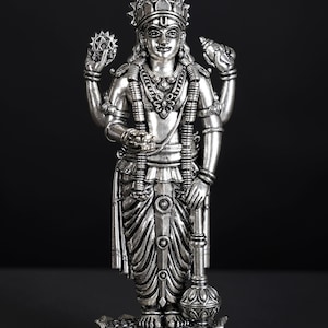 May include: A silver-colored statue of a deity with multiple arms, adorned with intricate jewelry and a crown. The figure stands on a lotus flower base, holding various symbolic objects. The statue is set against a dark background.