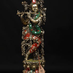 May include: A detailed brass statue of a deity playing a flute, with a green and red beaded necklace and red and gold clothing. The statue stands on a decorative base with colourful accents. The tag reads "exotic india".