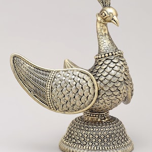May include: A brass-colored metal peacock figurine featuring intricate scale-like engravings on the body and base, with a textured wing design. The bird stands upright with a crest on its head and a detailed collar around its neck.